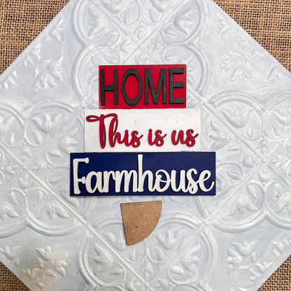 The Word Stacks - Painted sign by Janet's Craft Corner features "HOME" in red, "This is us" in red script, and "Farmhouse" in white script on a blue background with a small tan semicircle beneath the text on a white embossed tile.