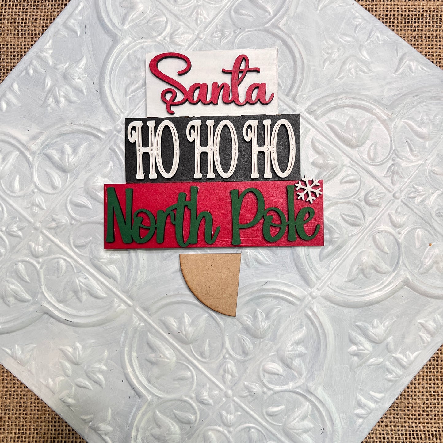 The Word Stacks - Painted by Janet's Craft Corner is a decorative sign featuring layered text—“Santa,” “HO HO HO,” and “North Pole”—with a snowflake, displayed on a textured white tile background.