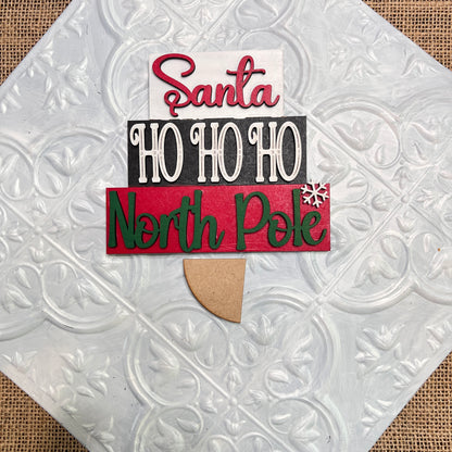 The Word Stacks - Painted by Janet's Craft Corner is a decorative sign featuring layered text—“Santa,” “HO HO HO,” and “North Pole”—with a snowflake, displayed on a textured white tile background.