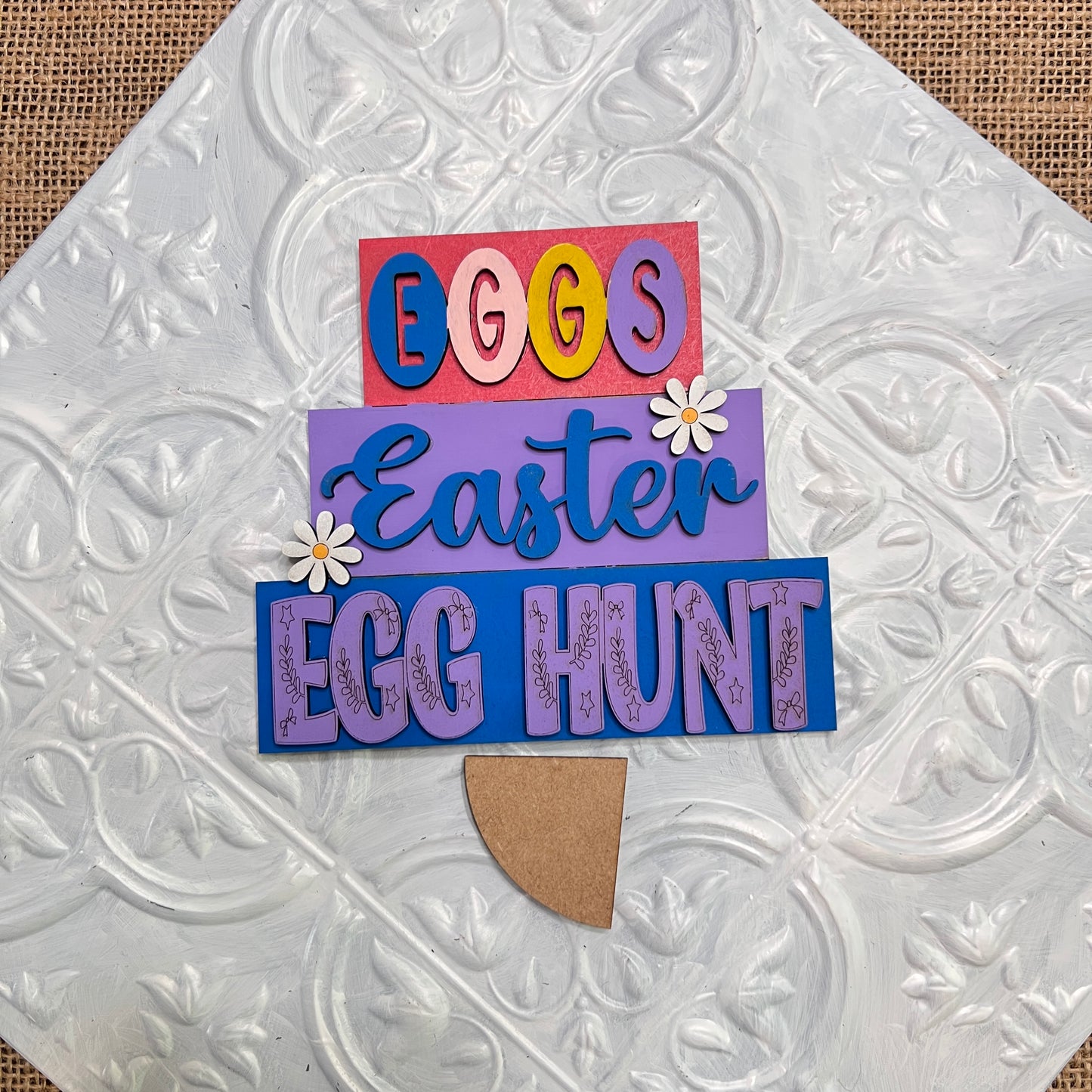 The Word Stacks - Painted by Janet's Craft Corner features stacked “EGGS,” “Easter,” and “EGG HUNT” lettering with daisies on a textured white background, designed to resemble a decorated Easter egg.