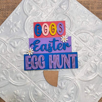 The Word Stacks - Painted by Janet's Craft Corner features stacked “EGGS,” “Easter,” and “EGG HUNT” lettering with daisies on a textured white background, designed to resemble a decorated Easter egg.