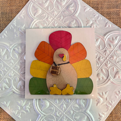A handmade turkey decor from Janet's Craft Corner features colorful red, orange, and yellow paper tail feathers and a wooden body with a "Gobble Gobble" sign, displayed on 6 inch squares.