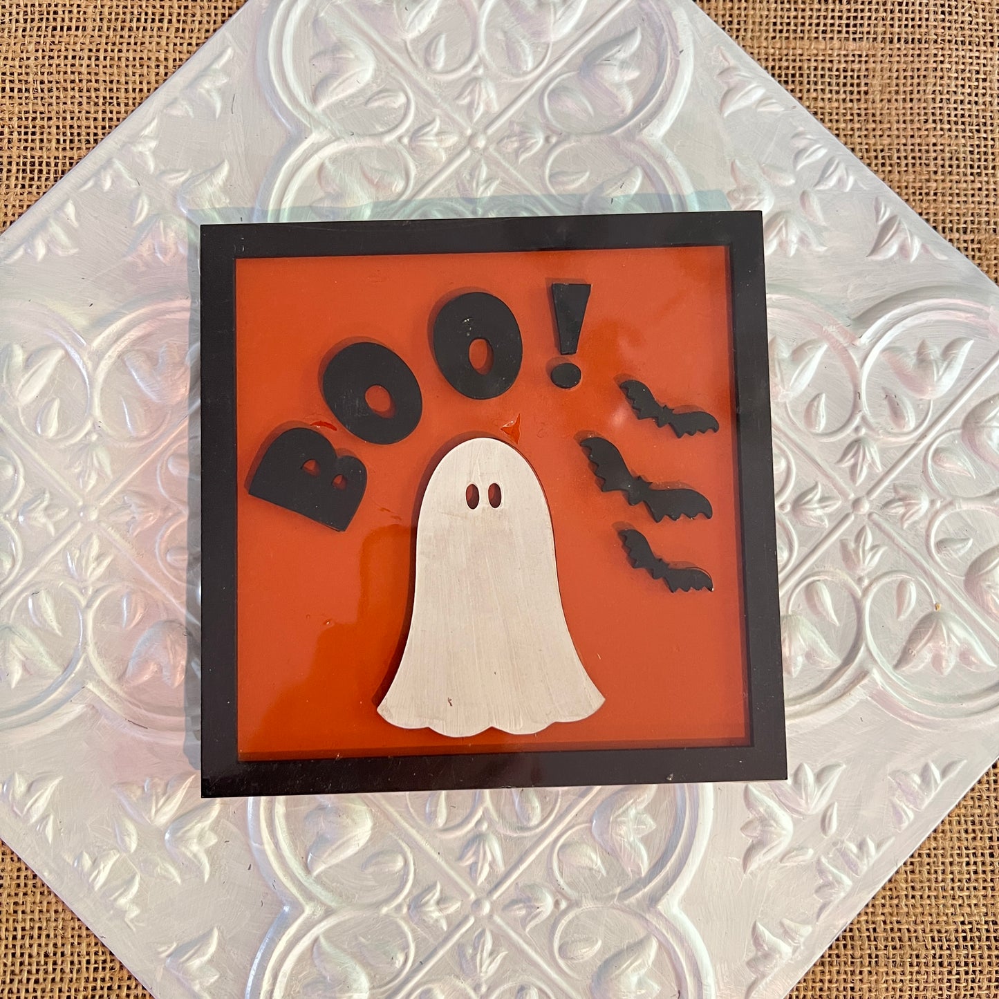 A 6-inch square Halloween sign from Janet's Craft Corner displays a white ghost, three black bats, and "BOO!" in black on an orange background with a black frame, set on a textured silver surface.