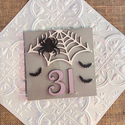 Janet's Craft Corner 6 inch square features a spider on a web, three bats, and the number “31” on a gray background with textured white tile and burlap underneath—perfect for Halloween decorating.