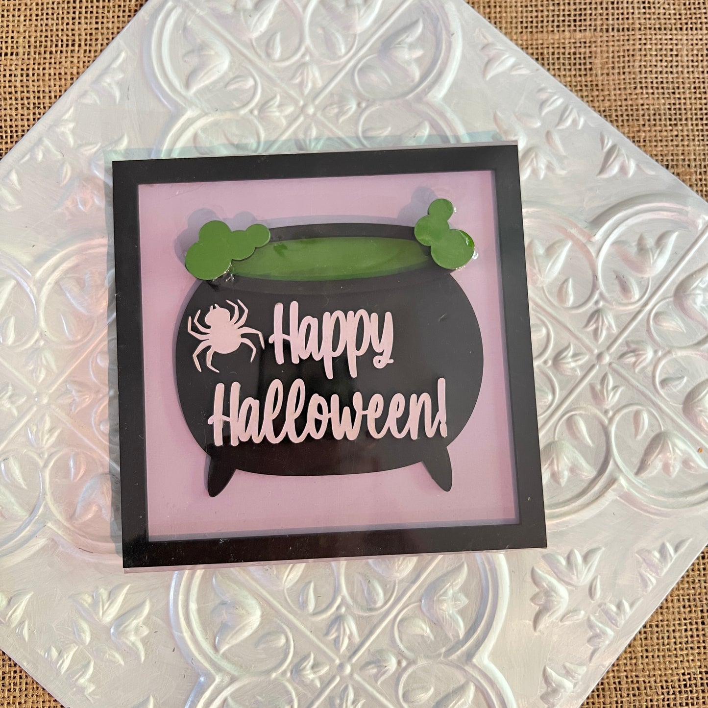 A 6-inch square sign from Janet's Craft Corner features a black cauldron, green bubbles, and a white spider with "Happy Halloween!" on a pink background, displayed on a patterned metallic surface.