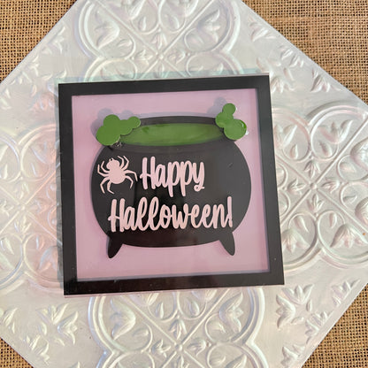 A 6-inch square sign from Janet's Craft Corner features a black cauldron, green bubbles, and a white spider with "Happy Halloween!" on a pink background, displayed on a patterned metallic surface.