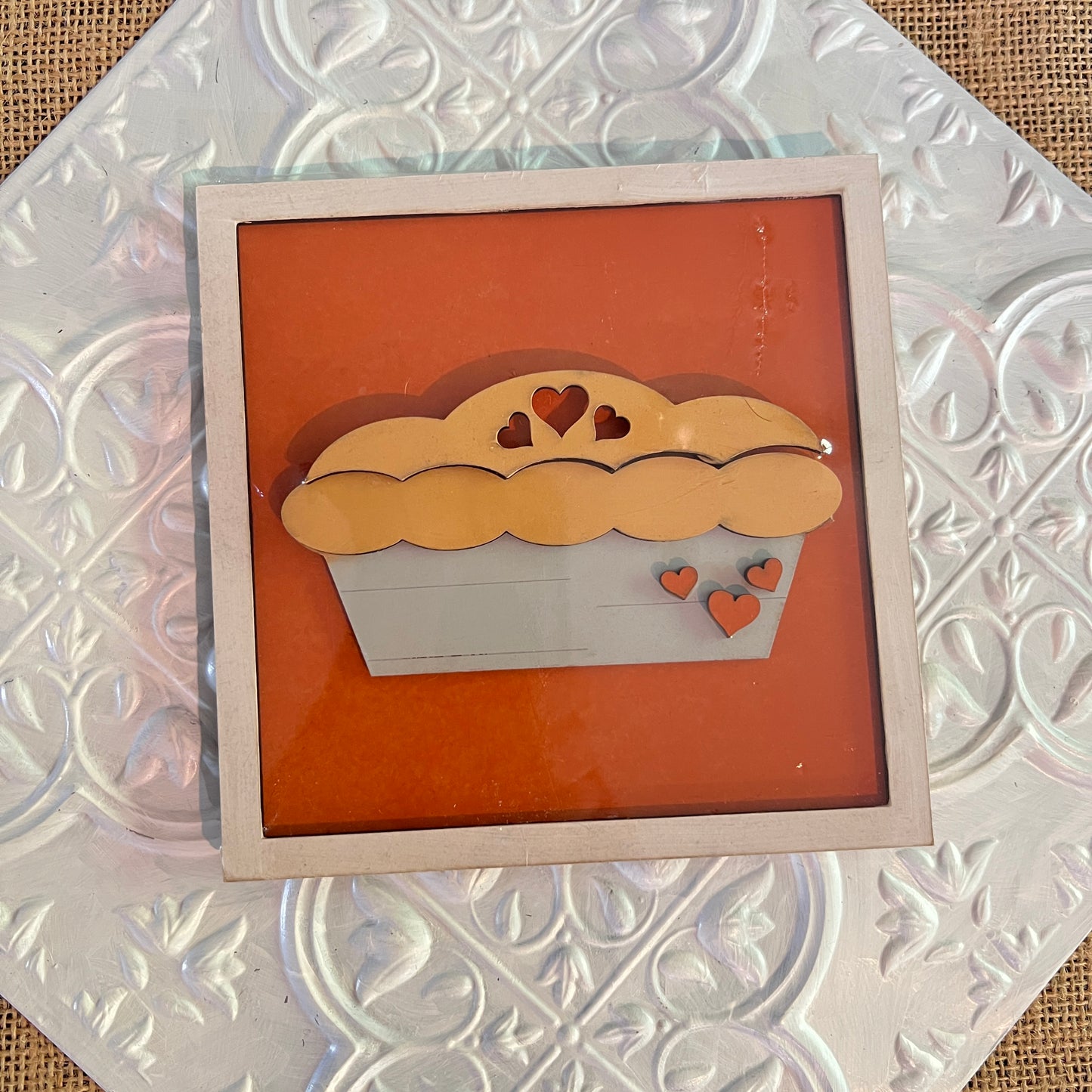 A framed artwork of a pie with heart-shaped cutouts rests against an orange background, displayed on a textured white "6 inch squares" tile by Janet's Craft Corner.