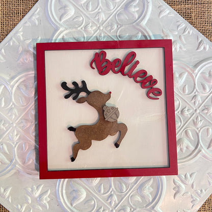 Janet's Craft Corner 6 inch square features a brown reindeer leaping with "Believe" in red above it, accented by a red frame on a white patterned background.