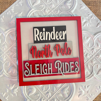 Janet's Craft Corner 6 inch squares: A decorative sign with "Reindeer" (white on black), "North Pole" (red script on gray), and "Sleigh Rides" (white/red on black) in a red frame on textured silver background.