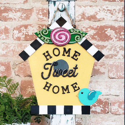 The Janet's Craft Corner "Home Tweet Home" birdhouse door hanger is a charming DIY decor piece, featuring yellow paint with black and white checkered edges, a pink flower, and a blue bird. It elegantly hangs on a wall, displaying "Home Tweet Home" in black letters.