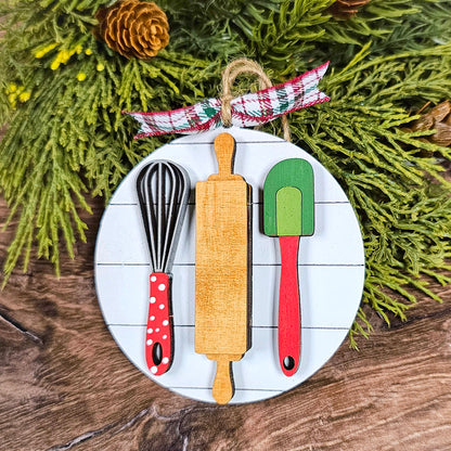 Introducing the Baking Spirits Bright Ornaments by Janet's Craft Corner! This individually painted festive ornament showcases wooden baking utensils, including a whisk, rolling pin, and spatula, each embellished with vibrant accents. Perfectly paired against a rustic backdrop enhanced by green foliage and pinecones, it is elegantly tied together with a plaid ribbon to brighten your holiday season.