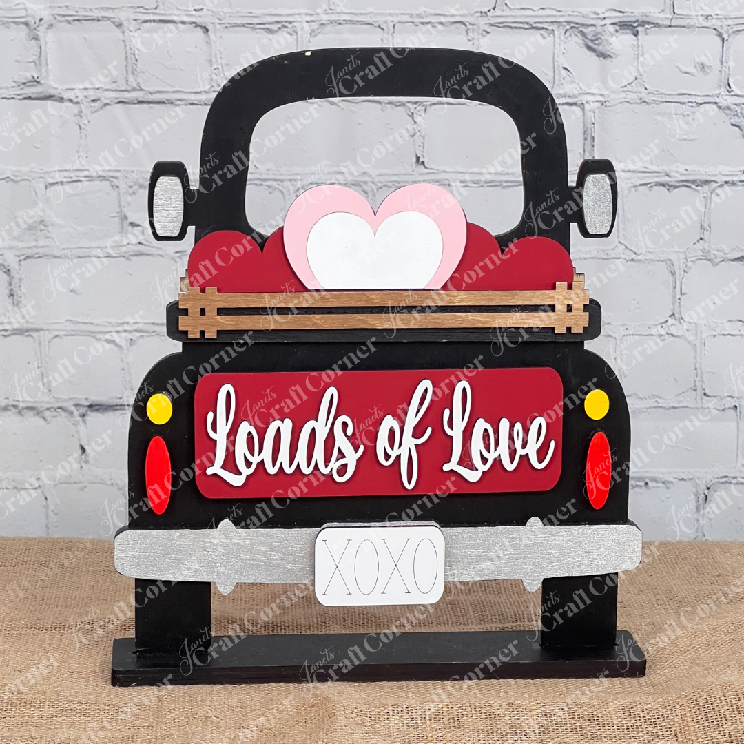 The Antique Truck Base with Insert DIY craft kit from Janet's Craft Corner includes a hand-painted truck with a wooden heart and "Loads of Love" decoration, plus an "XOXO" license plate. Ideal for interchangeable decor, it stands on burlap against a brick wall backdrop.