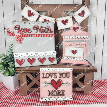 The Valentine Love Letters Decor Set by Janet's Craft Corner offers a charming DIY Craft Kit for home décor, featuring wooden items with signs like "Love Notes" and "Love You More" in pink, red, and white, displayed on a checked tablecloth with a small potted plant.