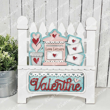 The Love Letters Interchangeable Insert from Janet's Craft Corner is a decorative wooden piece featuring Valentines-themed items like hearts and envelopes with pastel words "Love Letters" and "Valentine," set against a white picket fence, complete with a small potted plant for charm.