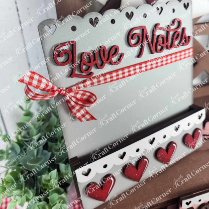 The Valentine Love Letters Decor Set by Janet's Craft Corner features a decorative card holder adorned with "Love Notes," red and white gingham ribbon, and heart patterns. Small red and white hearts embellish the bottom edge, while a background plant adds personalized charm.