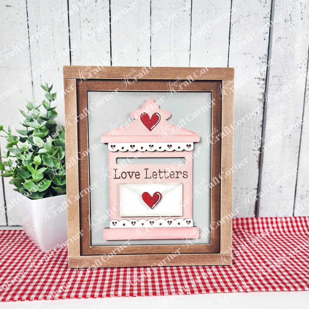 Janet's Craft Corner offers the "Love Letters Mailbox Interchangeable Sign," a DIY seasonal decor craft kit with a pink mailbox, two red hearts, and "Love Letters" wording against a white wooden background. It includes a potted plant and red checkered cloth. Set of 1.