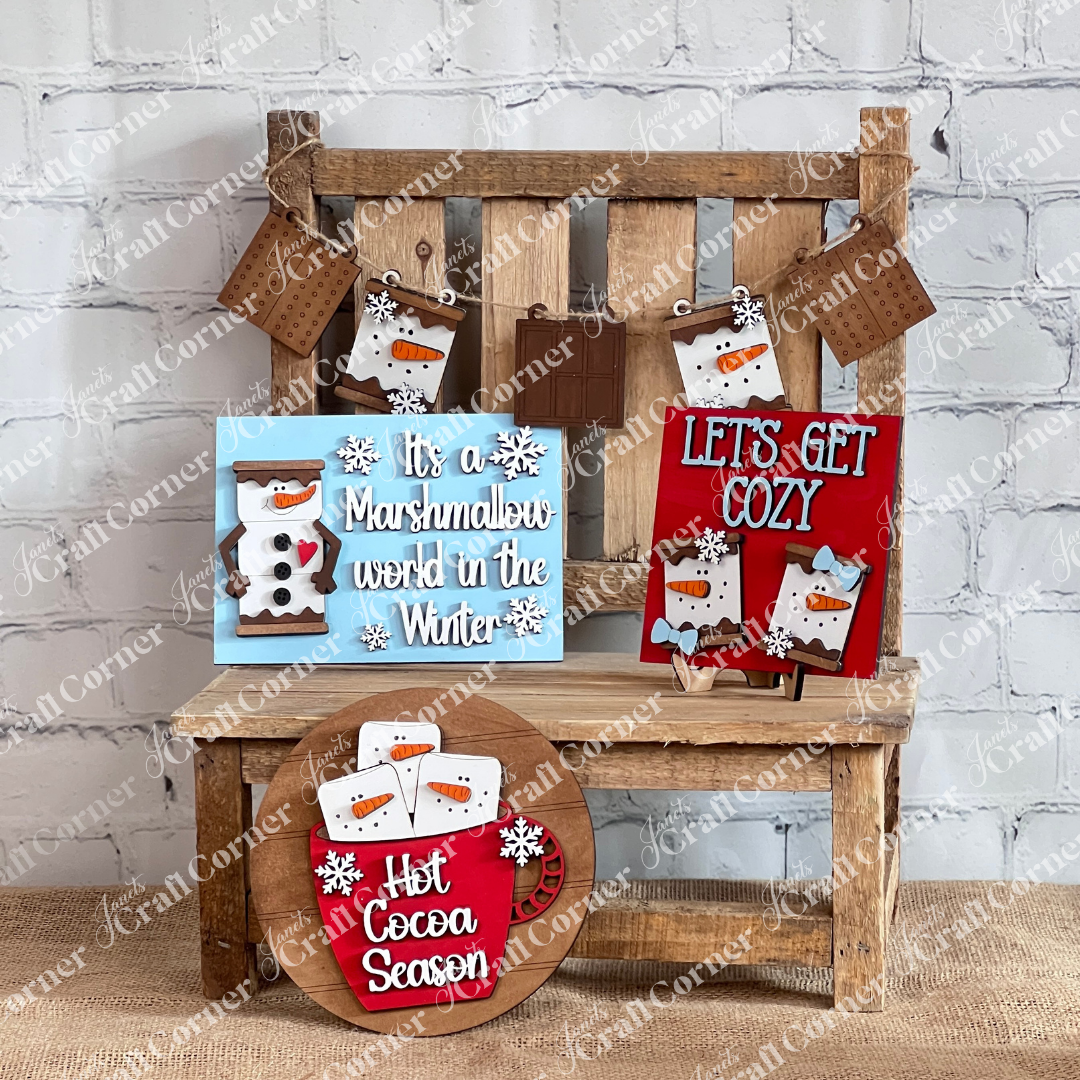 A rustic wooden bench showcases winter-themed décor from Janet's Craft Corner: the Marshmallow Snowman Smores Decor Set, which includes a blue sign with a Marshmallow Snowman and the phrase "It's a Marshmallow world in the Winter," alongside a red sign featuring gingerbread men with "LET'S GET COZY," and a round sign displaying mugs with "Hot Cocoa Season." Tags hang above.