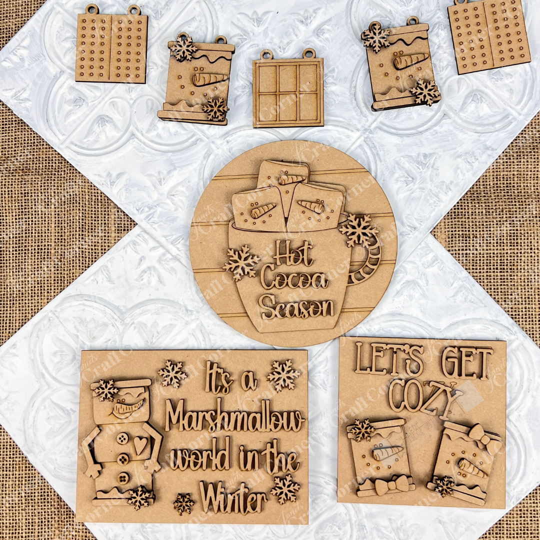 Top view of wooden winter-themed ornaments on a textured background. Featuring elements like hot cocoa mugs, snowflakes, and phrases such as "Hot Cocoa Season" and "It's a Marshmallow World in the Winter," the Marshmallow Snowman Smores Decor Set from Janet's Craft Corner invites you to create festive holiday decorations.