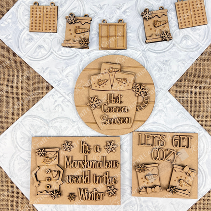 Top view of wooden winter-themed ornaments on a textured background. Featuring elements like hot cocoa mugs, snowflakes, and phrases such as "Hot Cocoa Season" and "It's a Marshmallow World in the Winter," the Marshmallow Snowman Smores Decor Set from Janet's Craft Corner invites you to create festive holiday decorations.