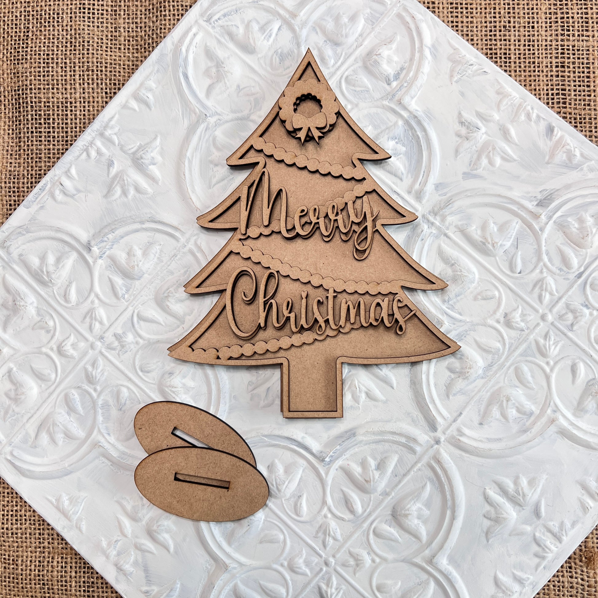A Standing Christmas Tree Shelf Sitter from Janet's Craft Corner features a wooden cutout engraved with "Merry Christmas" and is beautifully displayed on a textured white surface. Set on an oval wooden base and topped with a small wreath, this charming decoration is perfect for adding festive cheer to any shelf display.