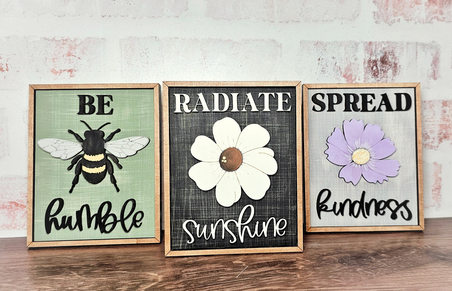 Janet's Craft Corner presents the Spread Kindness Farmhouse Sign series: "Be humble" with a bee, "Radiate sunshine" with a white flower, and "Spread kindness" with a purple flower. These framed signs on wood risers add charm against a faded brick wall in small spaces.