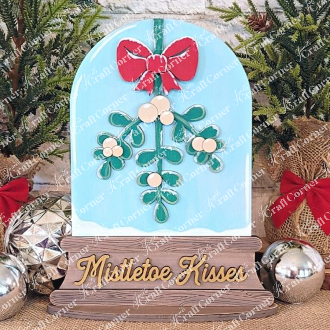 Janet's Craft Corner presents the "Mistletoe Kisses Snowglobe," an artistic decorative piece for home décor, featuring a mistletoe design with a red bow against a light blue background, adorned with festive elements like red bows and shiny ornaments, all labeled in elegant gold script.