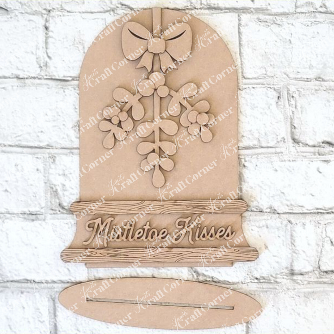 The "Mistletoe Kisses Snowglobe" by Janet's Craft Corner captures artistic expression with its mistletoe and bow design, along with the engraved words "Mistletoe Kisses." Ideal for home décor, it is beautifully set against a white brick background.