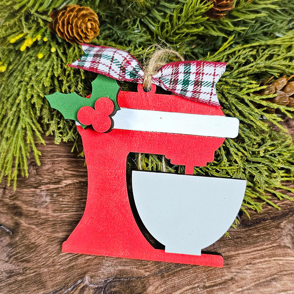 A hand-painted ornament, featuring a classic stand mixer with a bowl and adorned with holly and a plaid ribbon, hangs gracefully against a green pine background complete with pinecones. This charming piece from Janet's Craft Corner, known as the "Baking Spirits Bright Ornaments - Individual Painted," beautifully enhances your festive décor.