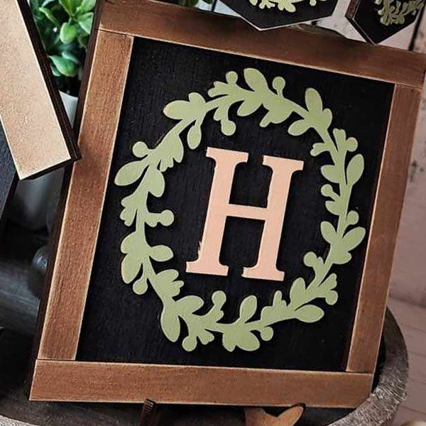 This monogram wall hanging from Janet's Craft Corner, ideal for home decor enthusiasts, features a decorative design with a wooden frame. At the center is a large letter "H," gracefully encircled by a laurel wreath motif in light green against a stylish black backdrop.