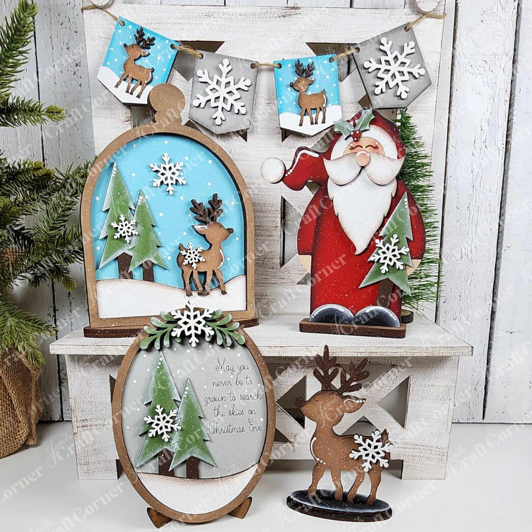 The Oh Deer Decor Set from Janet's Craft Corner features a festive display with hand-painted wooden decorations including Santa, reindeer, and snowflakes. Snowy trees and stars enhance the scene, accompanied by a charming sign that says, "May you never be too grown up to search the skies on Christmas Eve"—a true gem for home décor enthusiasts.