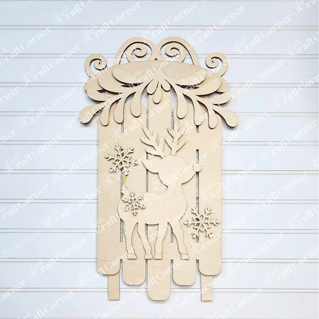 The Oh Deer Sled by Janet's Craft Corner features a hand-painted wooden laser-cut reindeer design, complete with antlers, snowflakes, and swirling elements against a picket fence background. Ideal for home décor, it is displayed on a white, horizontally lined surface.