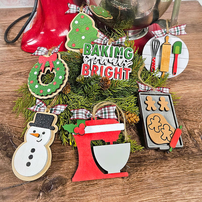 Individually painted ornaments from Janet's Craft Corner's Baking Spirits Bright collection are displayed on a table, featuring designs of a wreath, snowman, gingerbread cookies, and a red mixer. A sign says "Baking Spirits Bright." These unique ornaments are nestled among evergreen branches adorned with pinecones and festive ribbons.