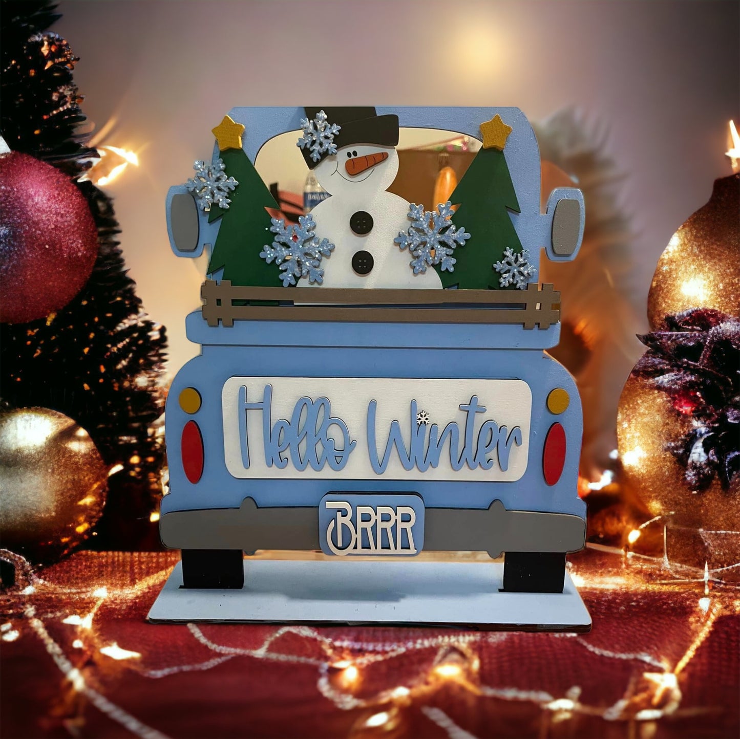 Create a festive holiday scene with Janet's Craft Corner's "Hello Winter" Interchangeable Insert - Painted. This charming decor piece features an antique blue truck adorned with "Hello Winter" on the back, surrounded by a delightful snowman, trees, snowflakes, and Christmas elements such as ornaments and string lights. Designed to offer interchangeable inserts for seasonal flair.