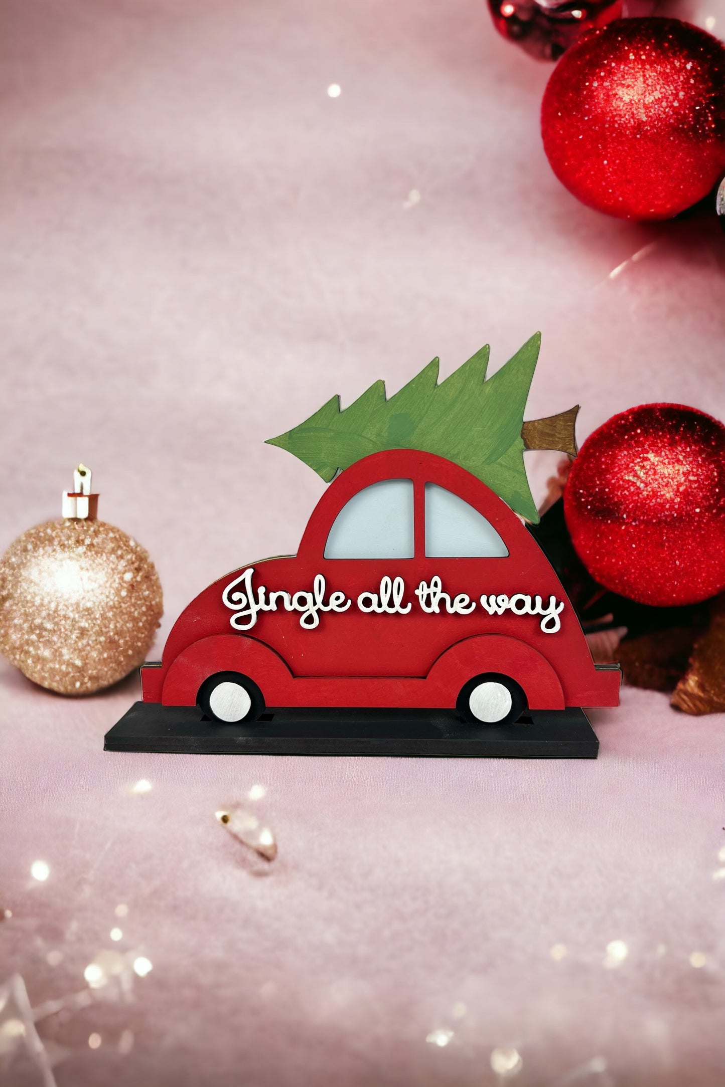 The "Jingle All the Way Volkswagen Shelf Sitter" by Janet's Craft Corner is a decorative red figurine with an evergreen tree on top, surrounded by red and gold Christmas baubles against a soft pink background with twinkling lights, adding yuletide cheer.