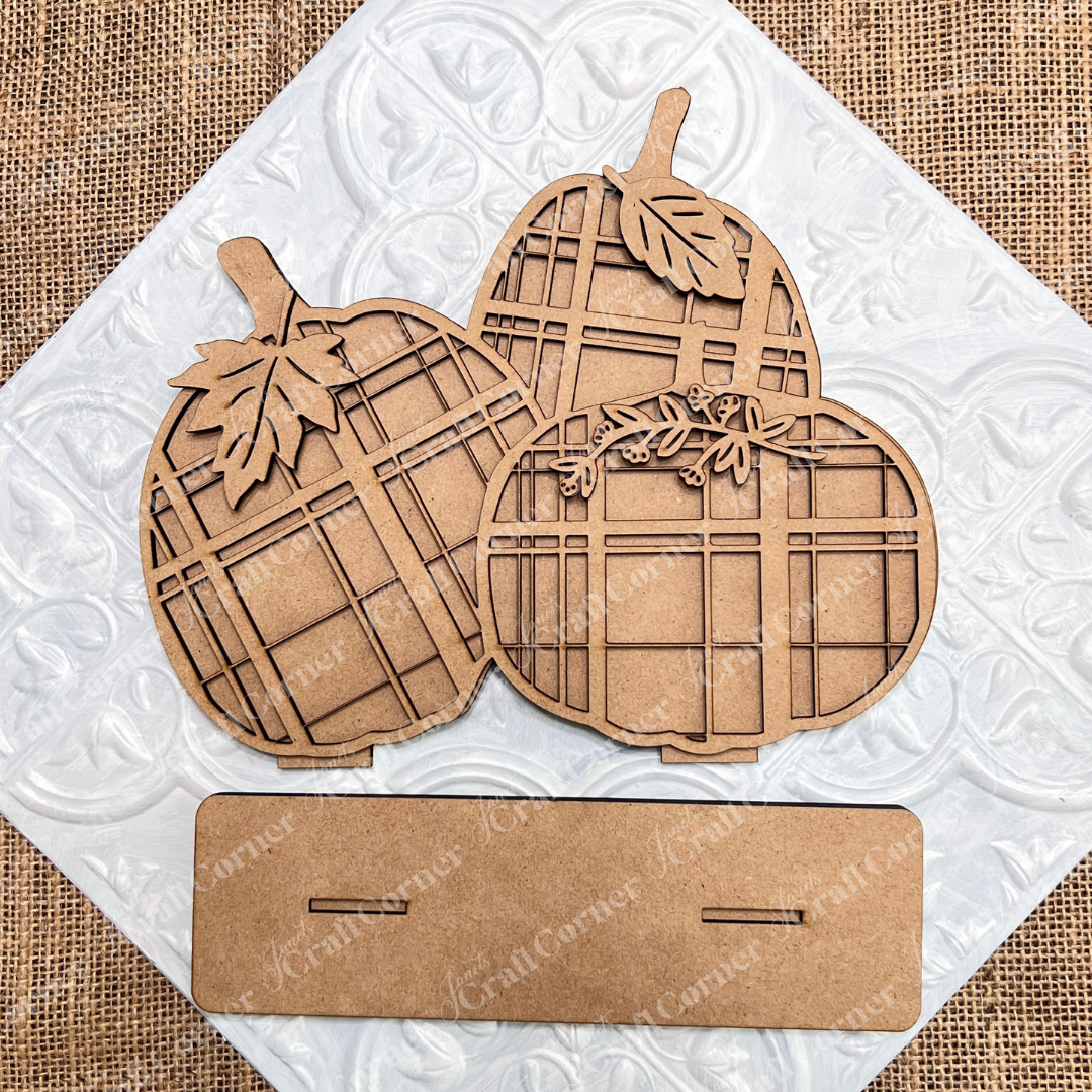 Three charming wooden pumpkin cutouts from Janet's Craft Corner, intricately etched with plaid patterns and leaves, are elegantly arranged on a textured white surface. This Plaid Pumpkins Shelf Sitter DIY home decor craft kit perfectly captures the essence of home décor. Below the pumpkins is a rectangular wooden stand featuring two slots for easy assembly.