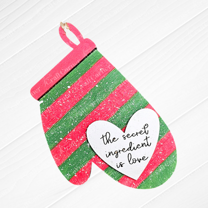 A delightful DIY craft kit from Janet's Craft Corner called "Baking Ornaments - Set of 2 | Early Access" showcases a striped mitten in red and green, accented with a heart-shaped cutout that says, "the secret ingredient is love." Ideal for adding charm to your home décor, this cute mitten is set against a white wooden background.