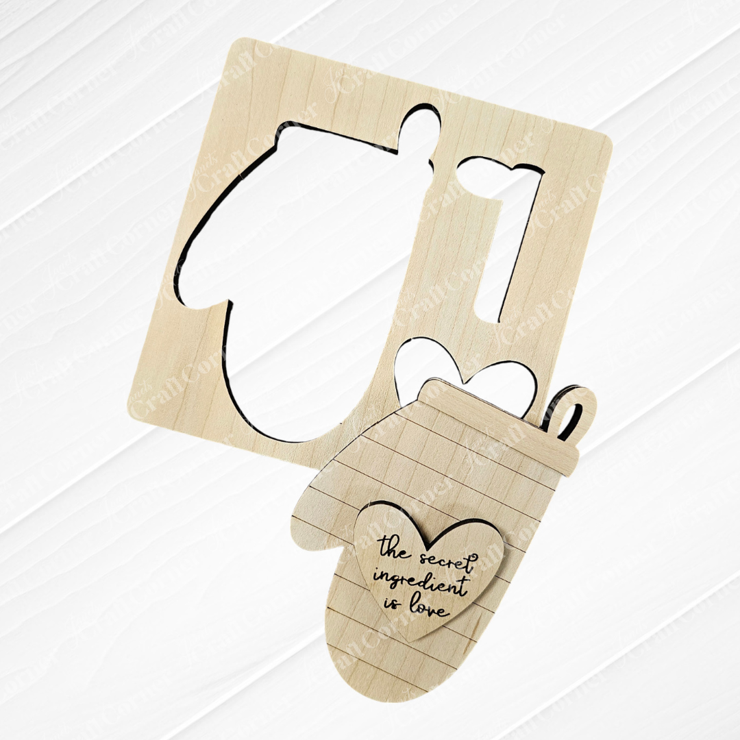 Discover the charming Baking Ornaments - Set of 2 by Janet's Craft Corner, featuring a wooden cutout of an oven mitt engraved with "the secret ingredient is love." Perfect for home décor, the mitt shape is partially removed from a square wood board. A subtle wood-textured white background enhances these delightful additions to your baking ornaments collection.