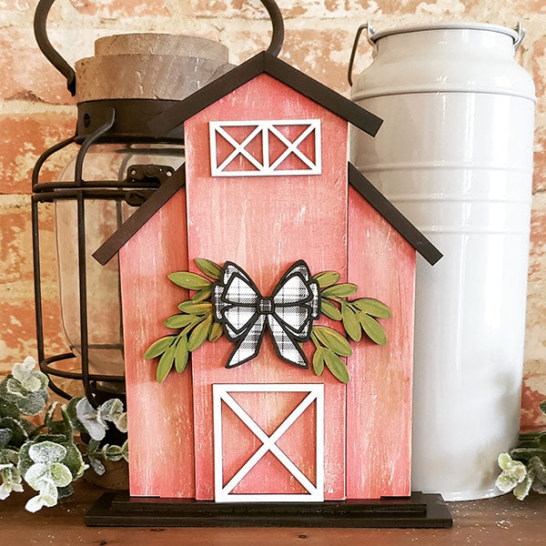 The Barn Shelf Sitter from Janet's Craft Corner is a decorative pink barn with white accents and a black-and-white bow, exuding rustic charm with its green foliage. Positioned against a rustic brick backdrop, it features a nearby black lantern and white metal container.
