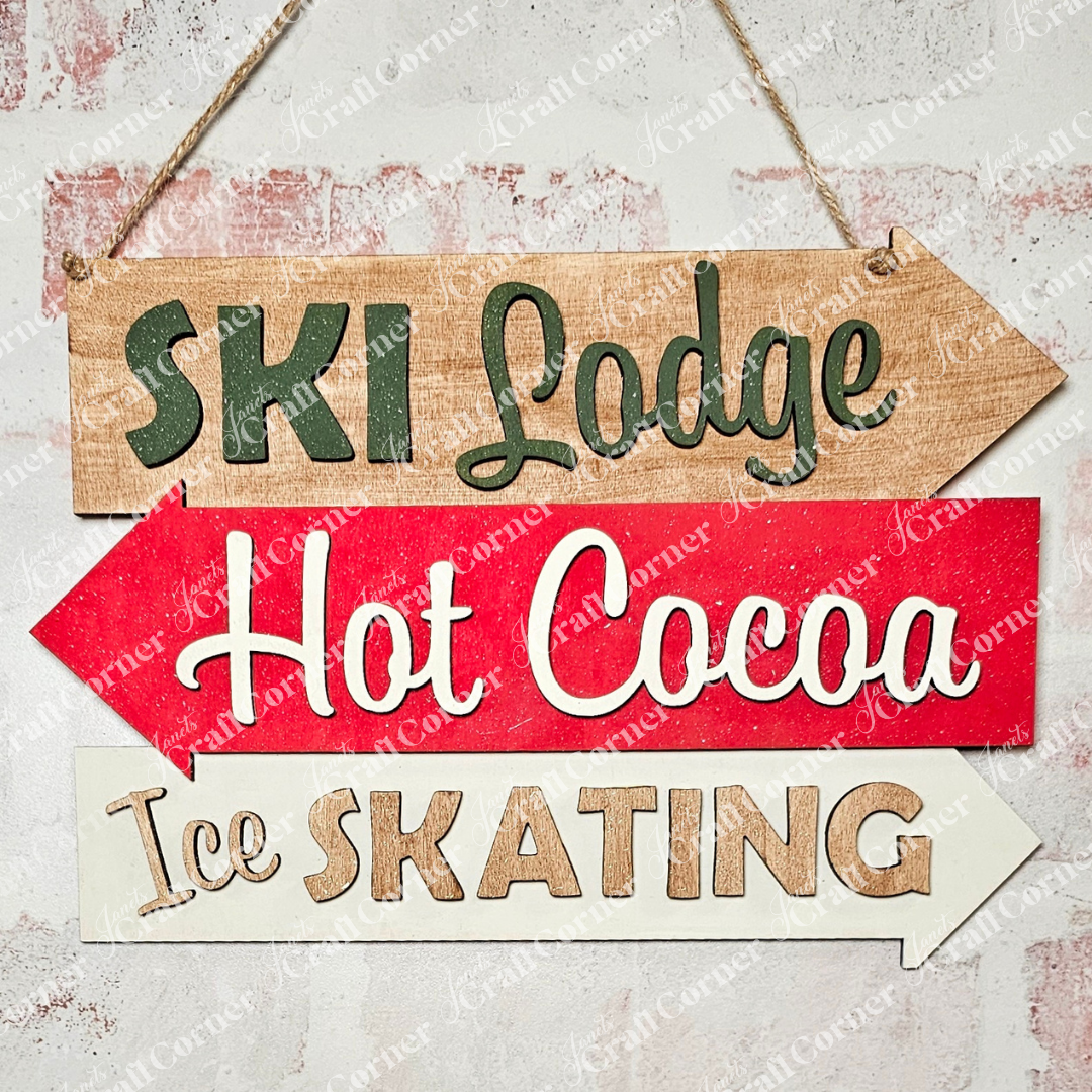 The Ski Lodge Directory Sign by Janet's Craft Corner is a charming wooden sign with hand-painted arrows: "Ski Lodge" in green, "Hot Cocoa" in red, and "Ice Skating" in white. Ideal for home décor or as a fun DIY craft kit, it hangs beautifully on a textured wall.