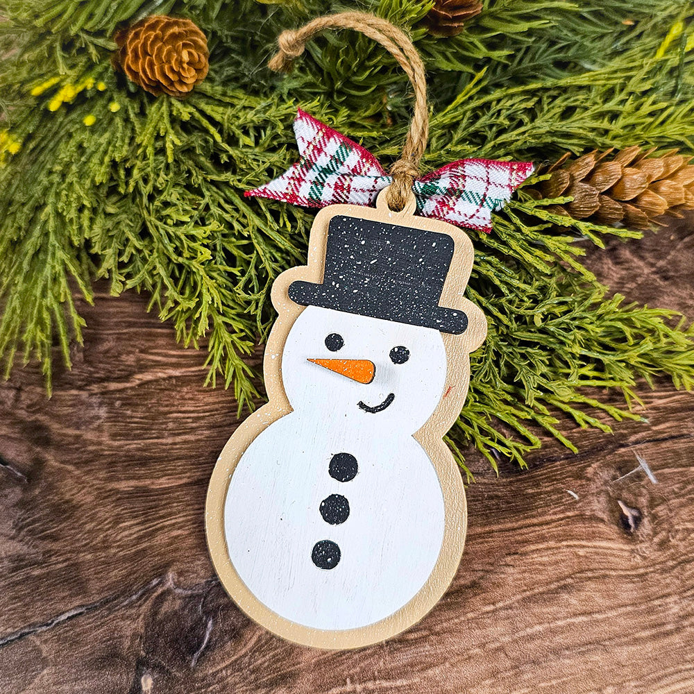 A handcrafted gingerbread cookie ornament with a black hat, orange nose, and black buttons hangs from a branch decorated with green foliage and pine cones. A plaid fabric bow adds charm to the Sugar Cookie Ornaments DIY Decoration Kit creation from Janet's Craft Corner, contrasting beautifully against the wooden background.