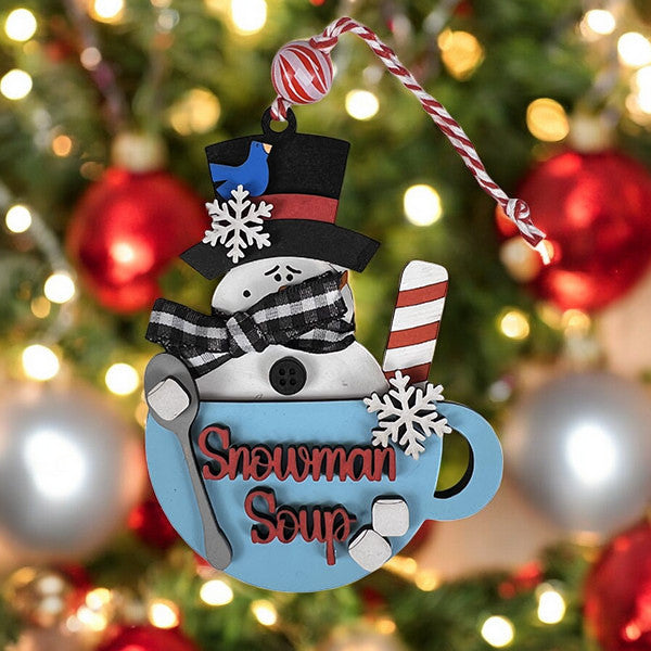 This festive Janet's Craft Corner Snowman Soup Ornament DIY craft kit features a charming snowman in a blue mug with "Snowman Soup" labeling. It sports a black top hat and scarf, with peppermint-style swirls, against blurred Christmas lights and red ornaments. Set of 1.