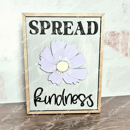 The "Spread Kindness" Farmhouse Sign by Janet's Craft Corner is ideal for tiered trays or small spaces. Featuring a light purple flower, it displays the word "SPREAD" at the top and "kindness" at the bottom in bold black on a light gray background, adorned with charming wood cut-outs.