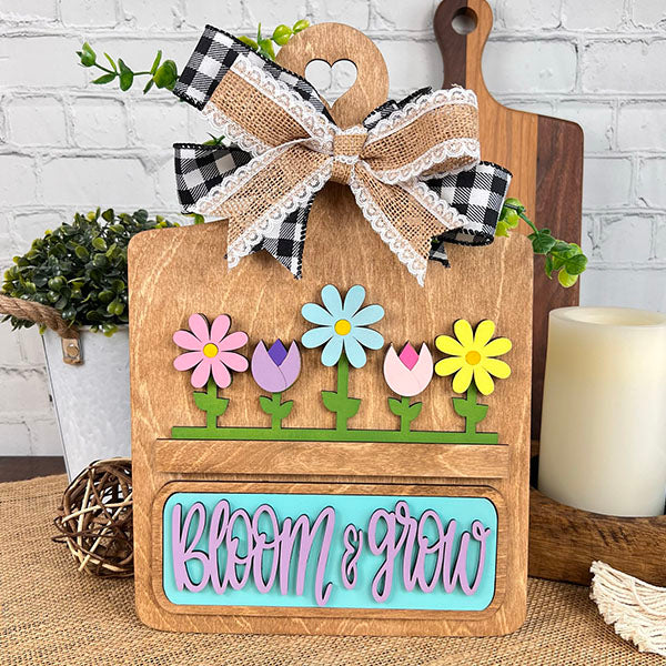 The Breadboard Interchangeable Base with Insert by Janet's Craft Corner is ideal for creative DIYers. Featuring colorful flower cutouts and "Bloom & Grow" in pink and blue, it includes a plaid and lace bow, complemented by items like plants, candles, and cutting boards against a brick wall.