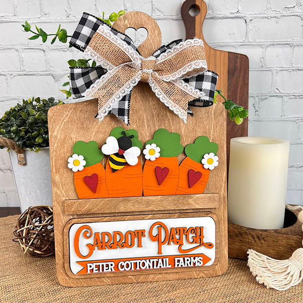 The Breadboard Interchangeable Base with Insert from Janet's Craft Corner is a delightful home decor piece featuring carrot cutouts with green tops and red hearts, a bee figure, and a checkered lace bow. It reads "Carrot Patch" and "Peter Cottontail Farms," perfect for adding rustic charm among plants and candles.