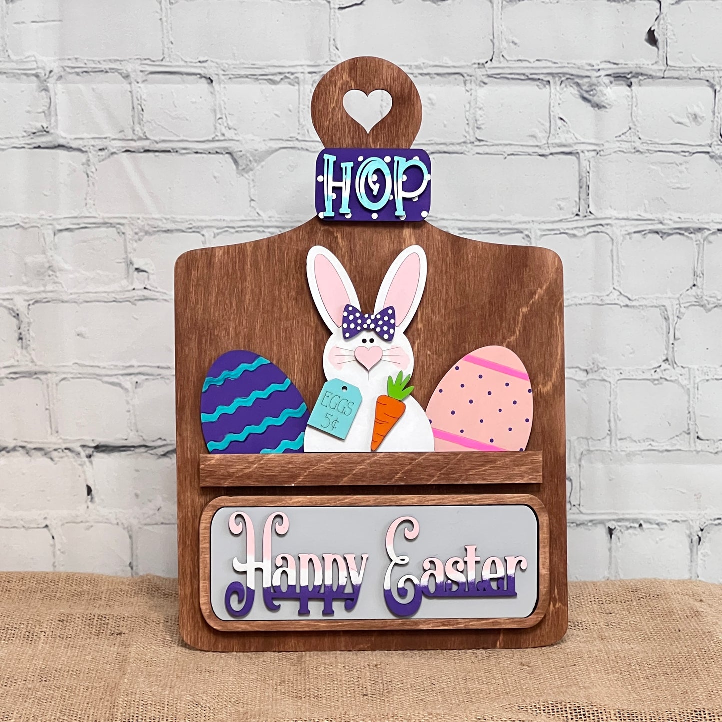 The Breadboard Interchangeable Base with Insert by Janet's Craft Corner is a DIY home decor kit featuring a wooden Easter sign: "HOP" at the top, a bunny with a carrot, surrounded by colorful eggs. Personalize it on a burlap-textured brick wall backdrop with "Happy Easter" charmingly at the bottom.