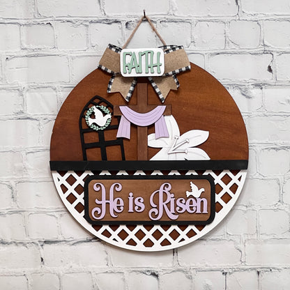 A round wooden decoration from Janet's Craft Corner hangs on a white brick wall, featuring "FAITH," a cross with purple drape, lilies, bold "He is Risen" text, and two doves. This interchangeable door hanger is part of a versatile home décor kit.