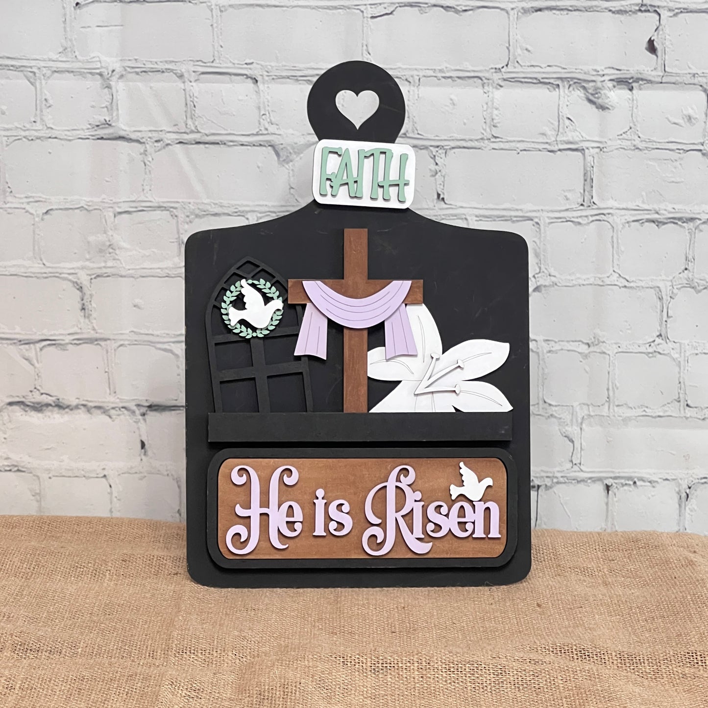 Janet's Craft Corner offers a DIY home decor kit featuring a breadboard base and insert with a cross draped in cloth, window silhouette with dove, and "He is Risen" in lilac text topped with "Faith," perfect for adding your personal touch against a white brick wall.