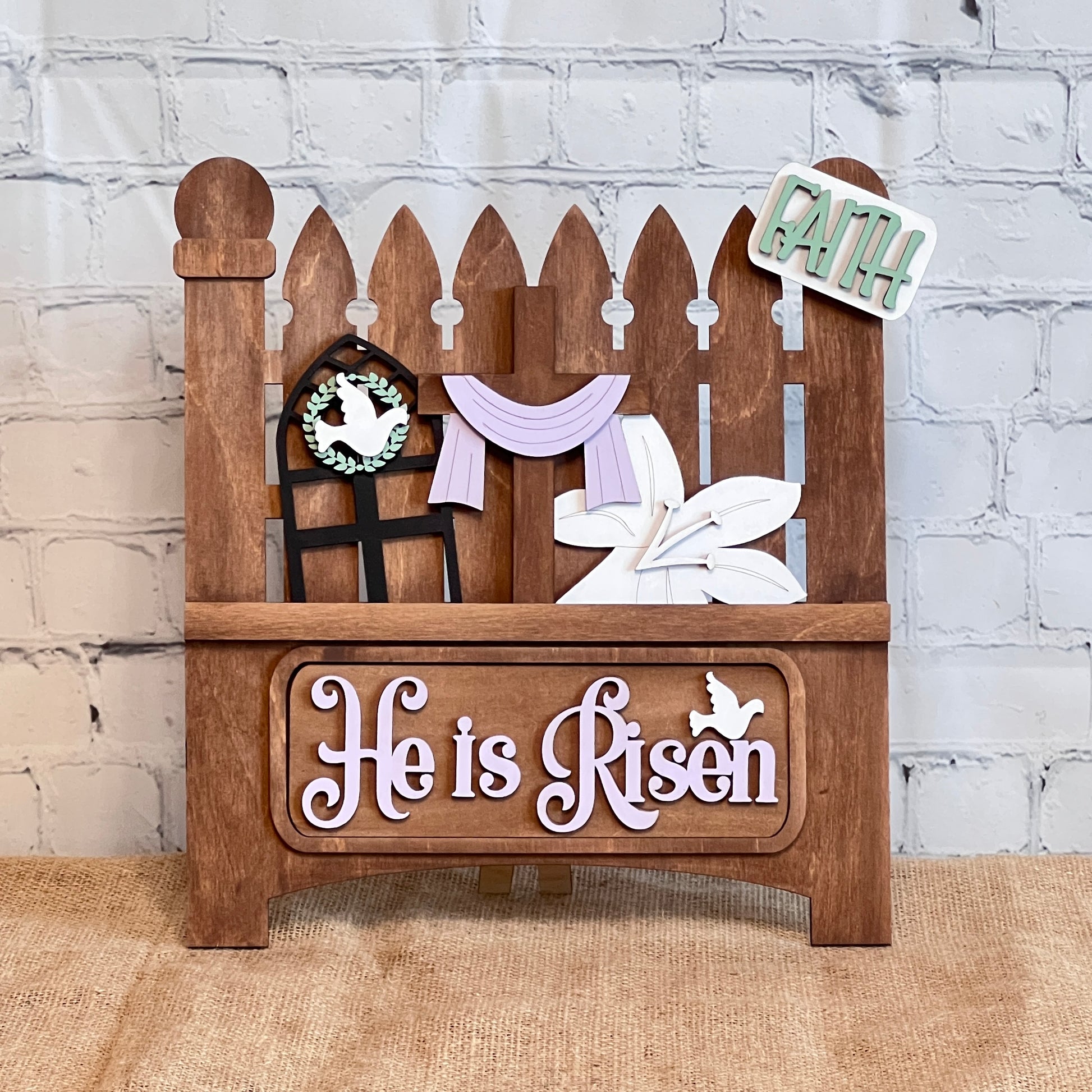 This delightful craft kit from Janet's Craft Corner features a Garden Fence design with a cross, purple cloth, white lily, "He is Risen" text, a small dove, and a "Faith" sign, all set against a light-colored brick wall backdrop. It includes 1 fence base and 1 insert.