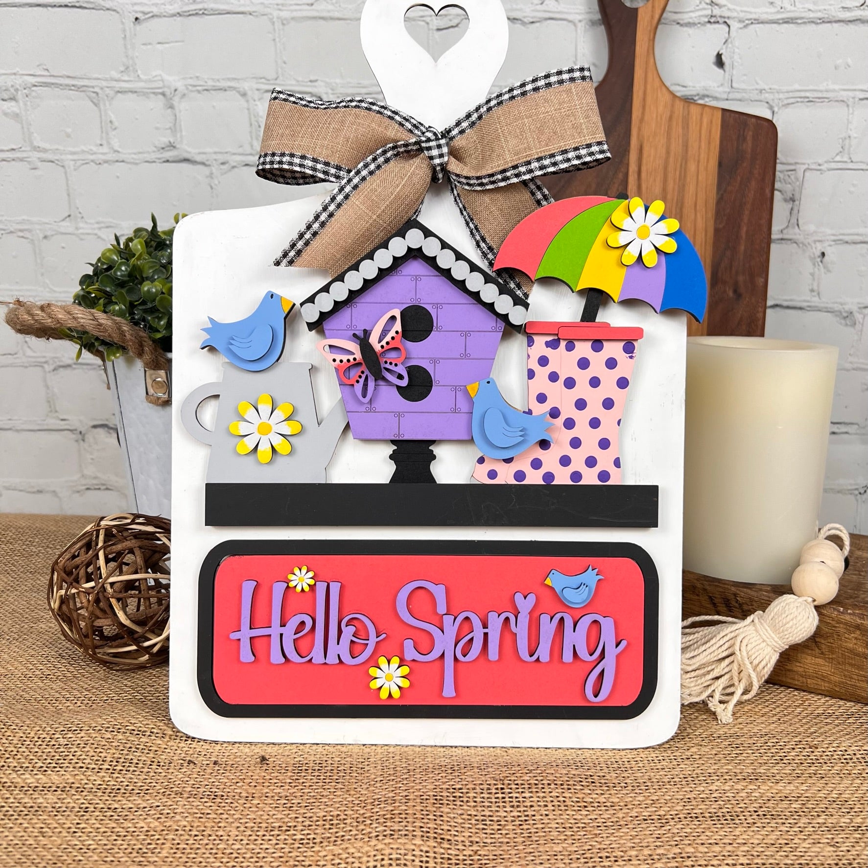 The whimsical Janet's Craft Corner DIY kit features a breadboard base and insert adorned with a birdhouse, umbrella, and flower-covered rain boots. A ribbon-tied board displays "Hello Spring." A candle and wicker balls complete this delightful home decor ensemble.