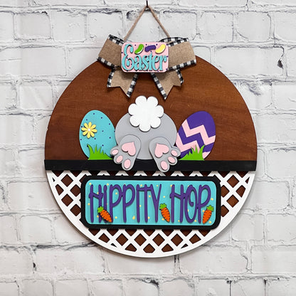 The DIY craft kit from Janet's Craft Corner features a bunny's backside with colorful eggs, a burlap "Easter" bow, and "Hippity Hop" with carrots. A white lattice borders the lower half against a brick wall background. The set includes 1 door hanger base and 1 insert.